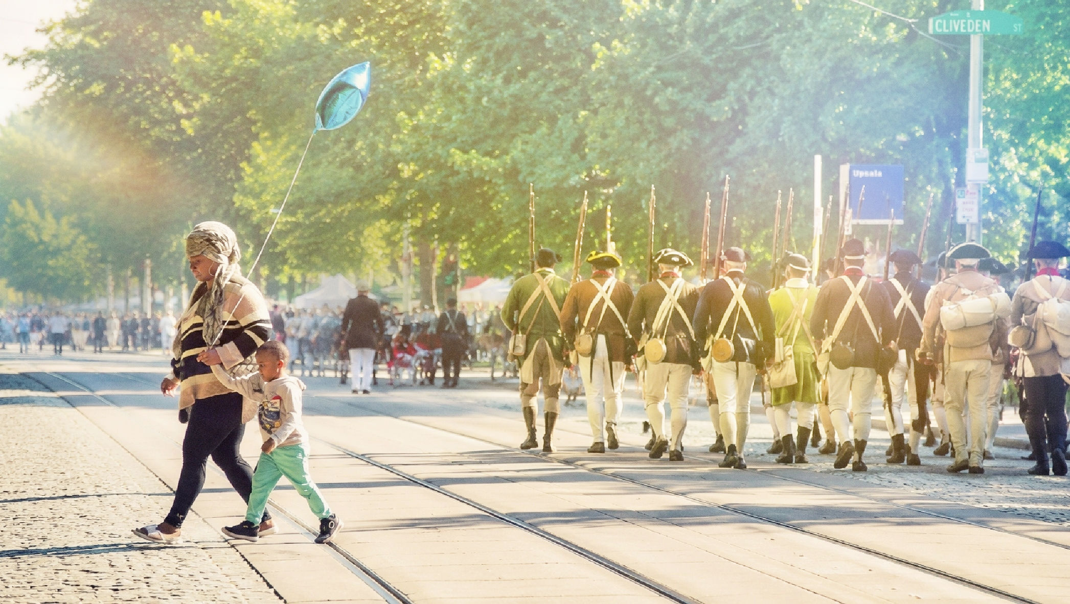 A woman and child with a blue balloon cross Germantown Avenue as Revolutionary War reenactors march near Upsala during the Revolutionary Germantown Festival.