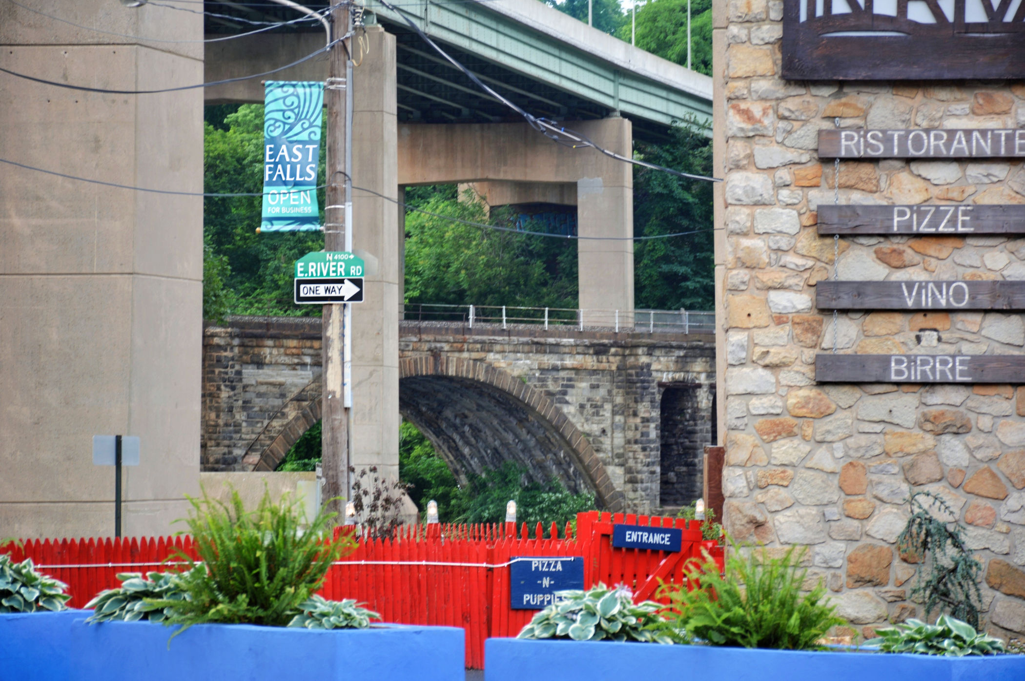 Colorful planters and red fencing at In Riva in East Falls, with the Twin Bridges and historic stone railroad arches rising behind the restaurant.