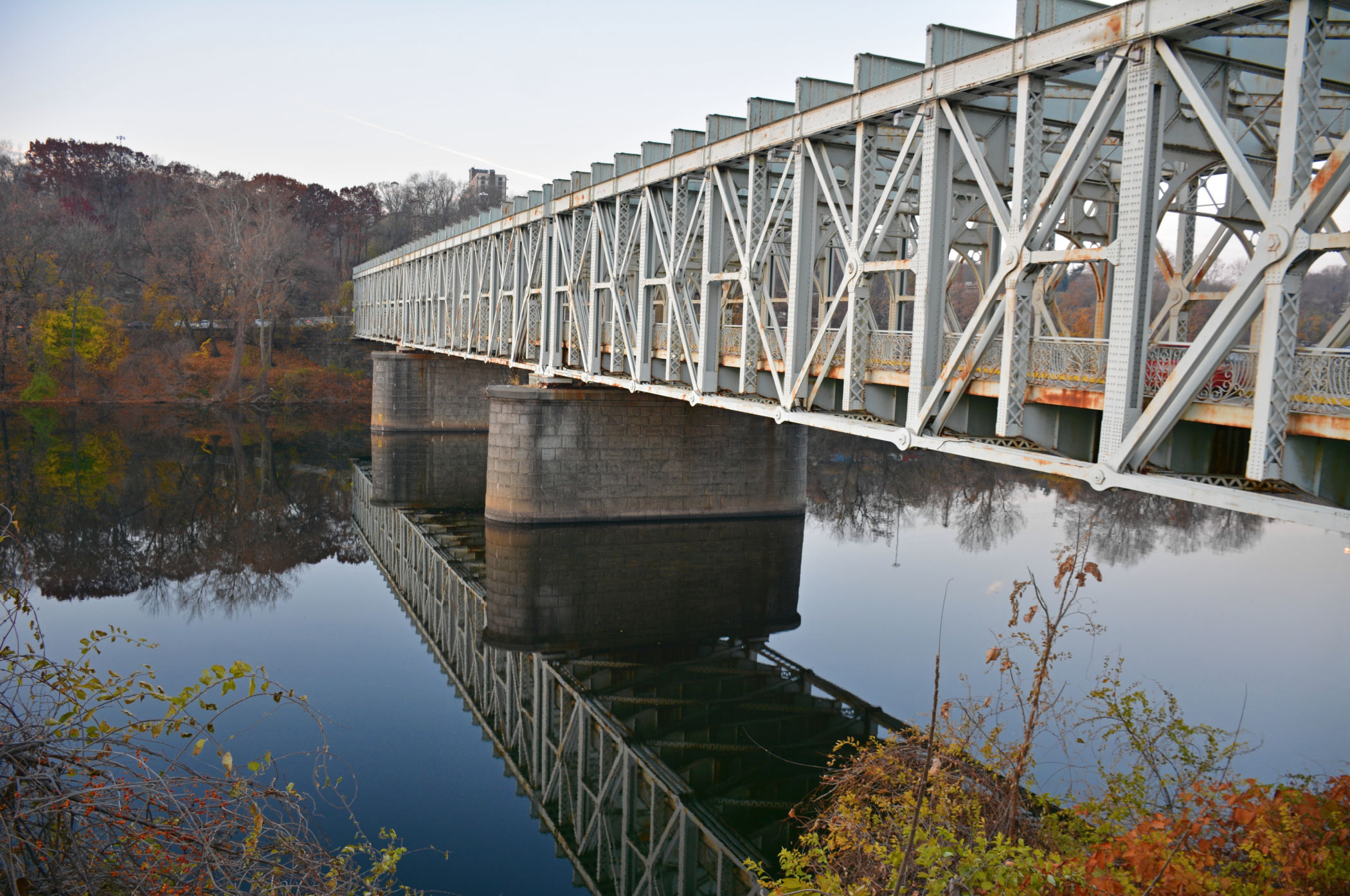 Angled view of the historic Falls Bridge in East Falls, showing its intricate steel framework and massive stone piers reflected in the Schuylkill River.