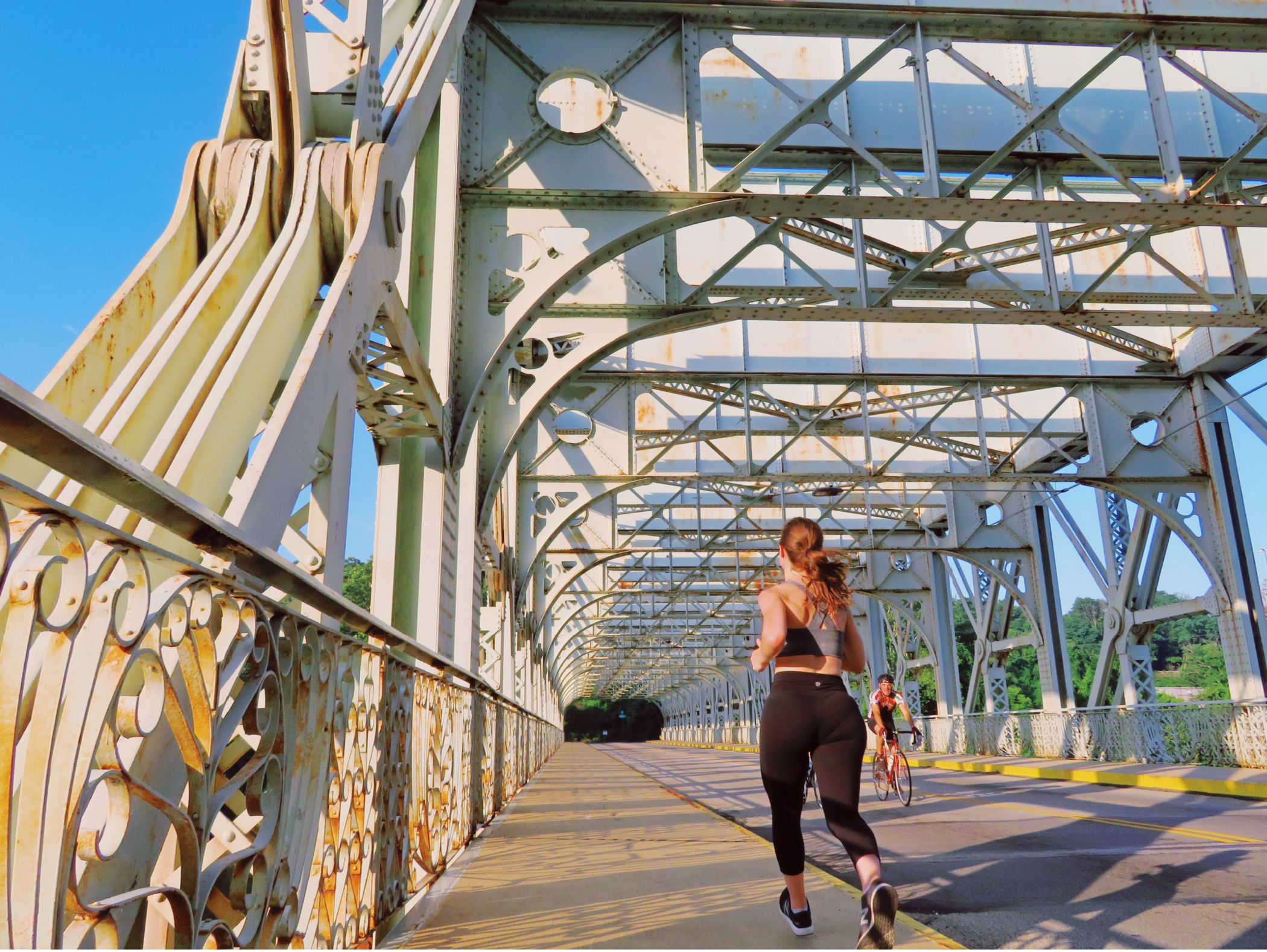 A runner and cyclist cross Philadelphia’s historic Falls Bridge in East Falls, surrounded by its intricate steel beams, geometric patterns and ornamental railings.