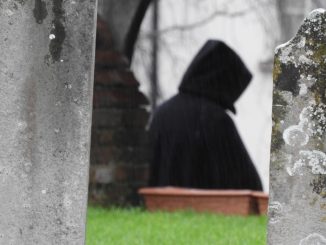 Hooded figure walking through a cemetery between weathered gravestones.