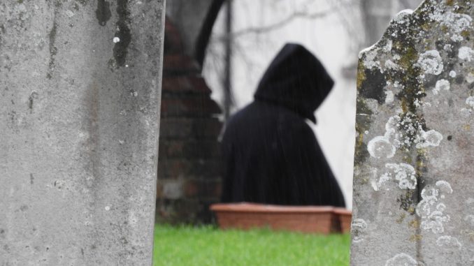 Hooded figure walking through a cemetery between weathered gravestones.
