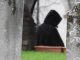Hooded figure walking through a cemetery between weathered gravestones.