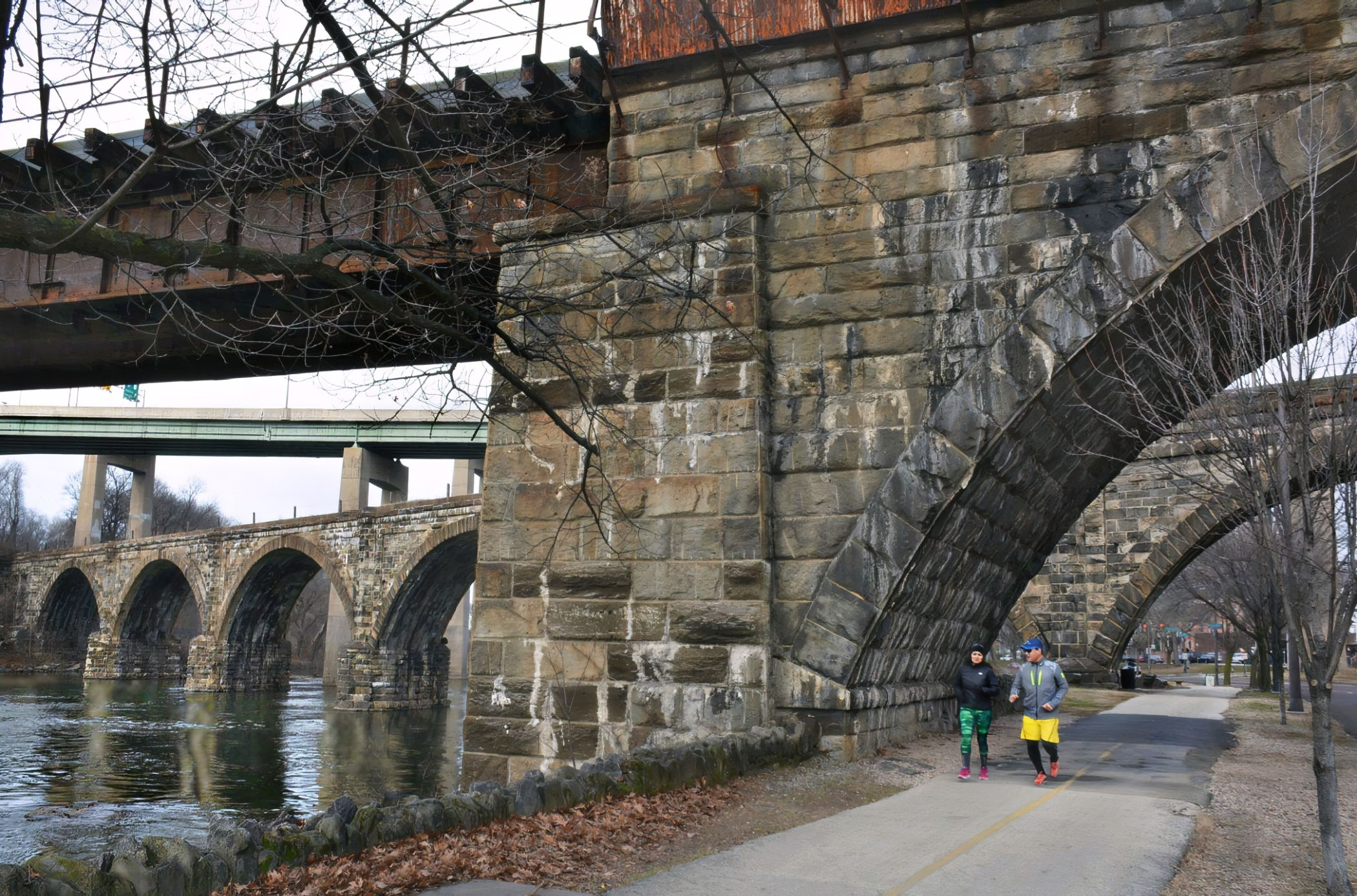 Two joggers pass beneath the massive stone arch of the Philadelphia and Reading Railroad Bridge near Kelly Drive in East Falls, with older railroad arches in the background.