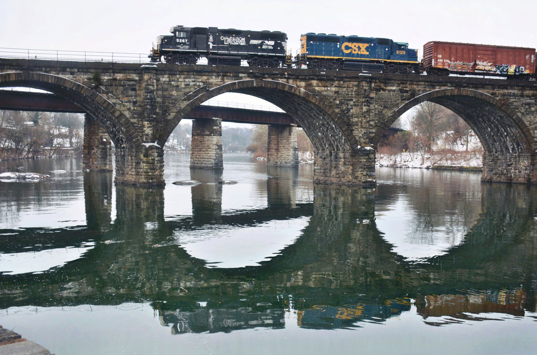 Freight train crosses the historic Reading Richmond Railroad Bridge in East Falls, with its stone arches reflected in the Schuylkill River.