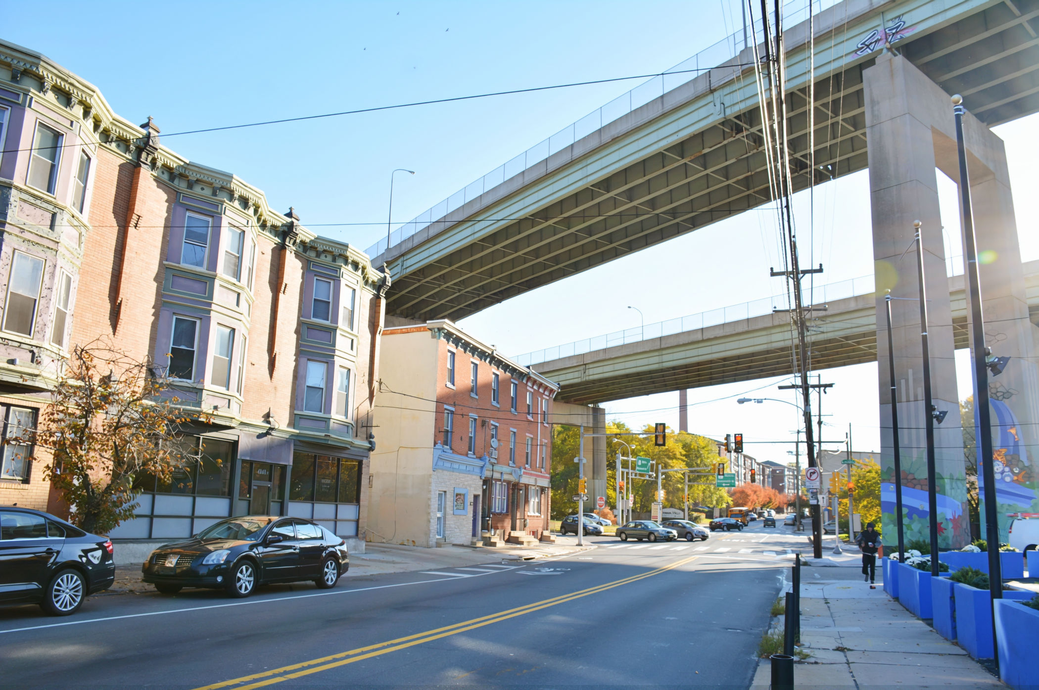 The Twin Bridges tower above brick rowhouses, storefronts and traffic along Ridge Avenue in East Falls, Philadelphia.
